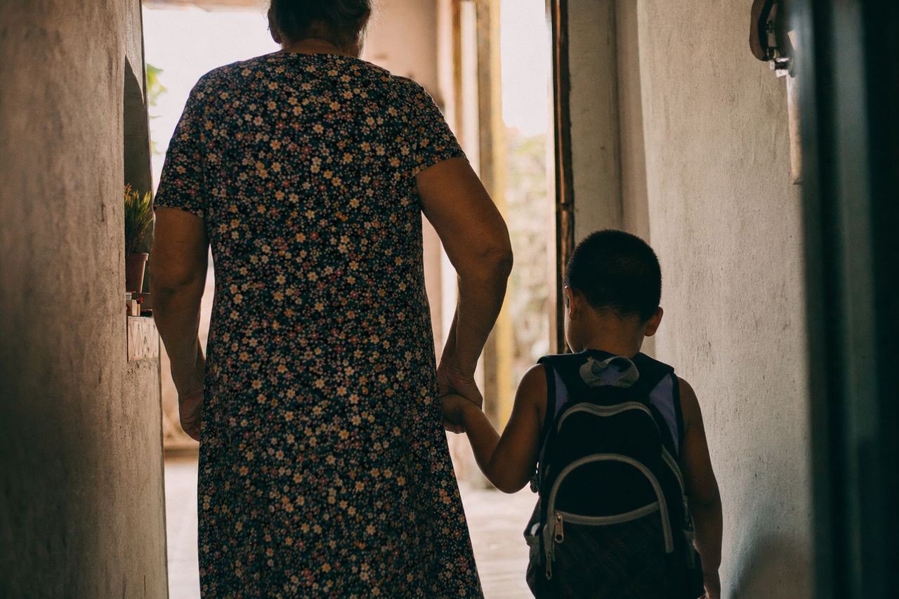 Avó e neto de mãos dadas, vistos de costas, caminhando pelo corredor de casa